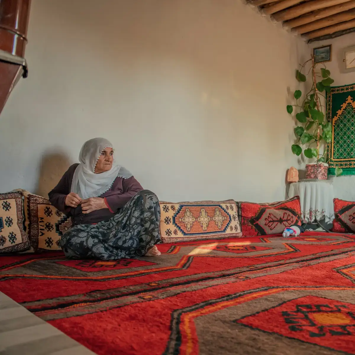 Mulher turca com vestes tradicionais, sentada no chão de sua casa, em cima de um tapete colorido estilo kilim
